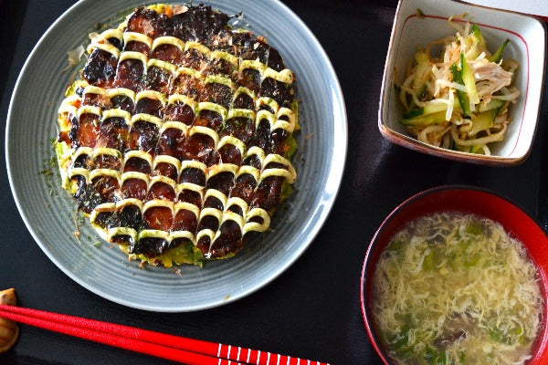 Okonomiyaki,egg soup, chicken and cucumber Asian salad are on the black serving tray.